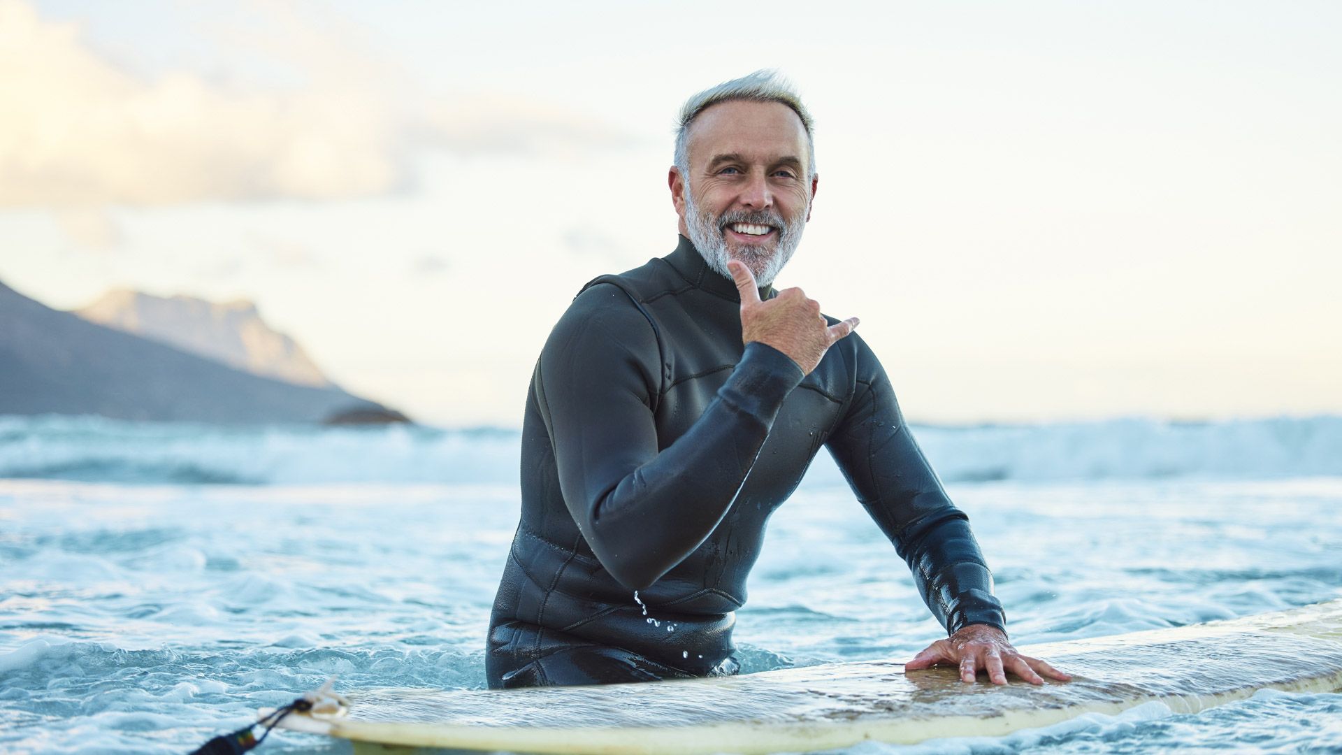Elderly man smiles while sitting on surfboard in the ocean with his thumb up as he looks into the camera