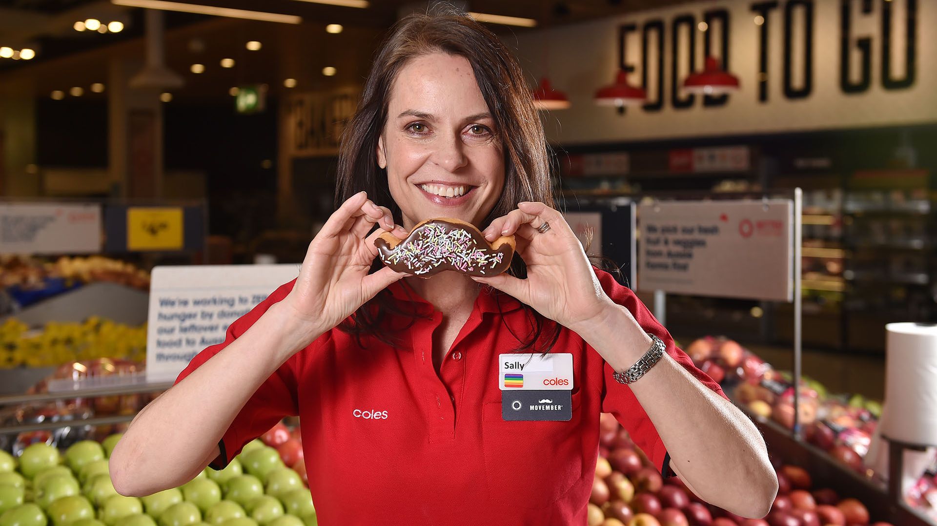 Photo of Coles employee smiling to camera, holding a moustache-shaped doughnut.