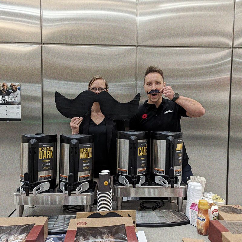 Photo of two people behind a coffee stand, holding an oversized Movember moustache.