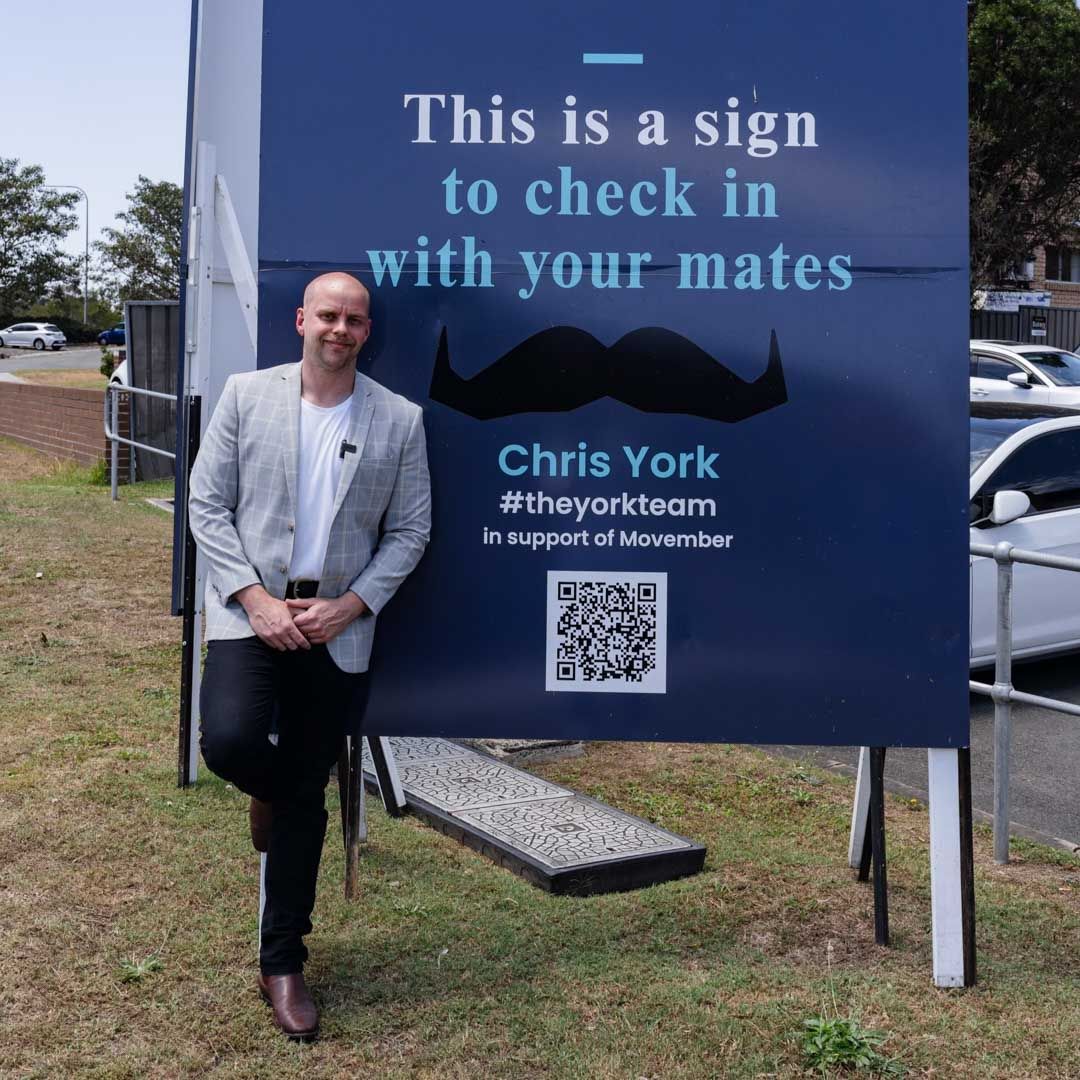 Photo of a man standing in front of a sign that promotes mental health and Movember.