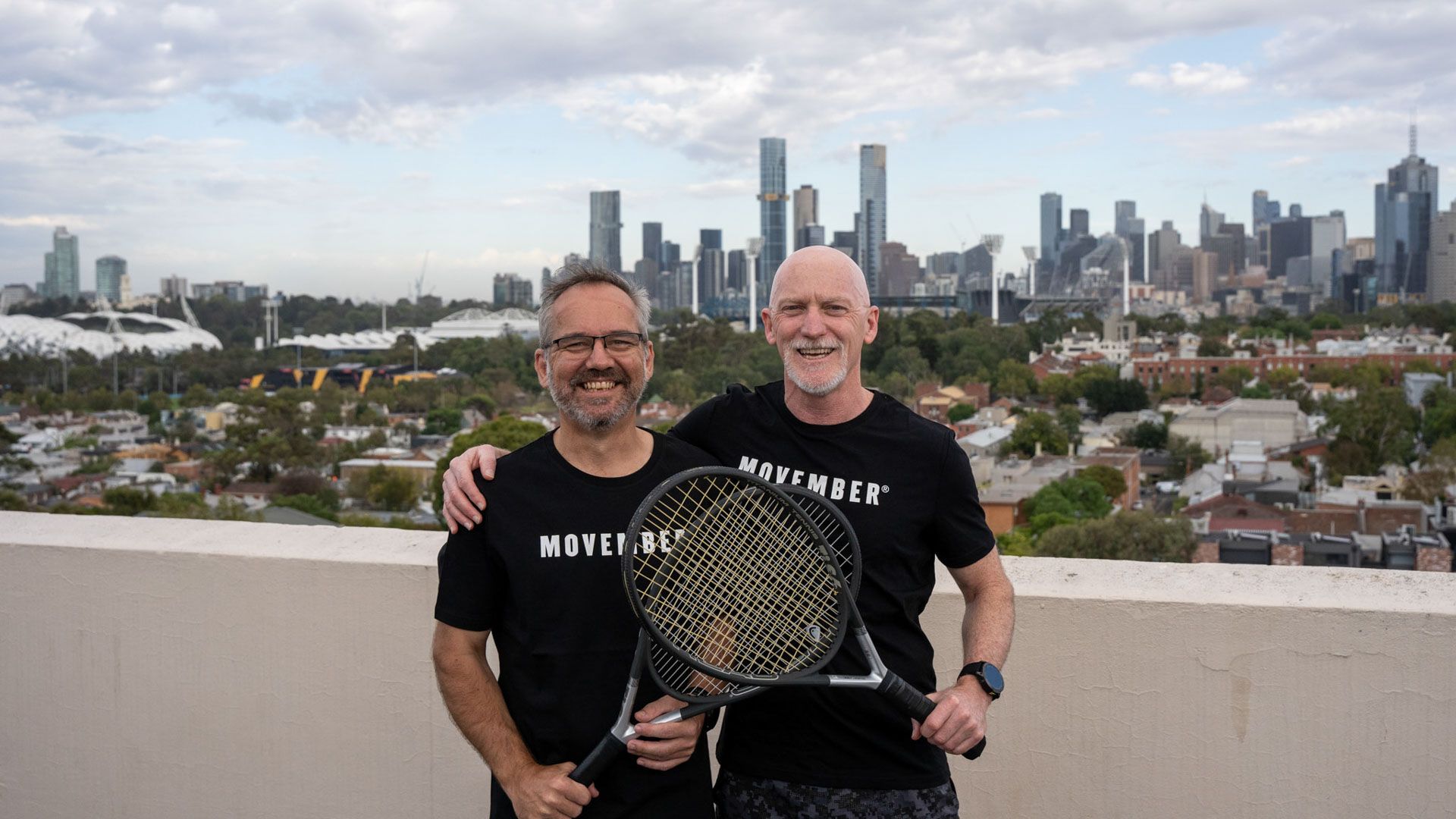 Two men holding tennis rackets, wearing Movember t-shirts, standing on rooftop with city scape in the background. 