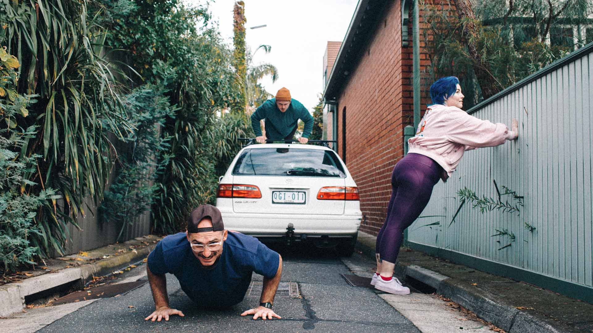 Three people performing push-ups in a suburban driveway.