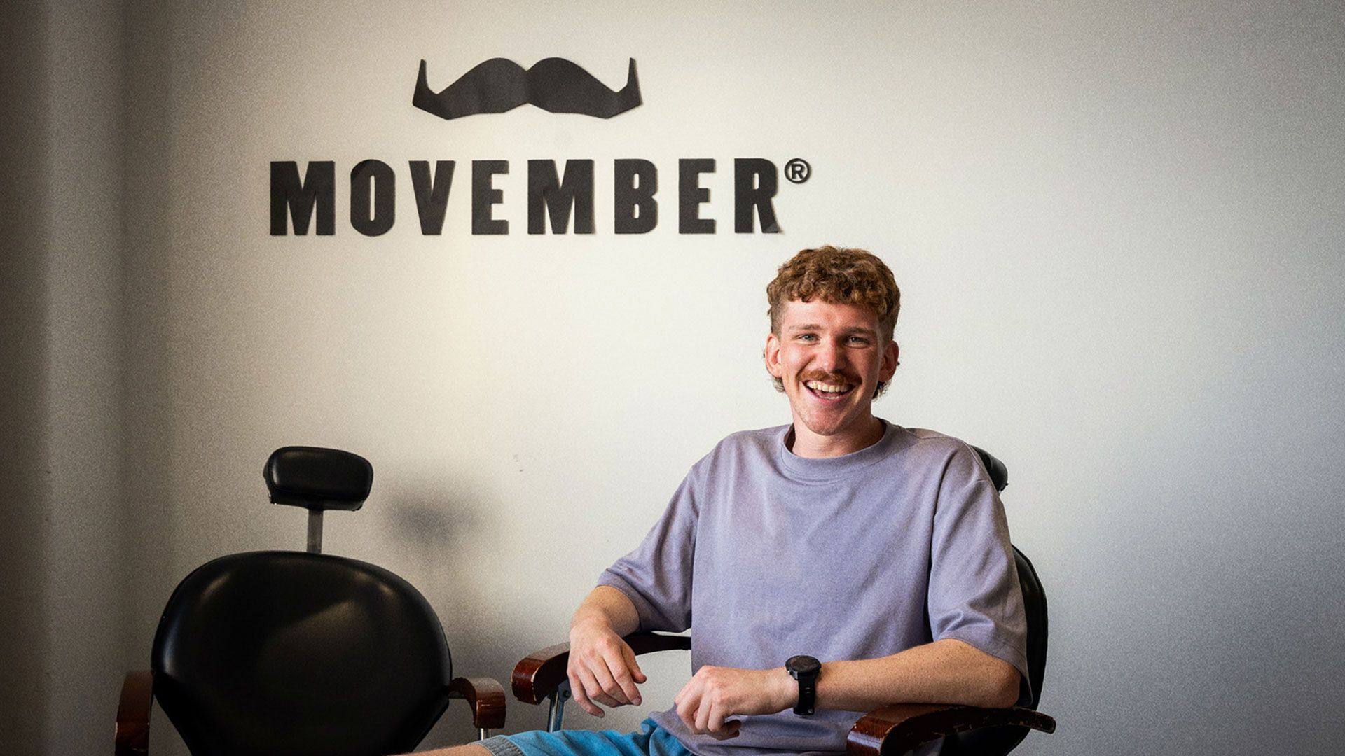 Young man sits in barber chair with Movember sign on background behind him. He smiles looking at the camera wearing a light purple t-shirt.