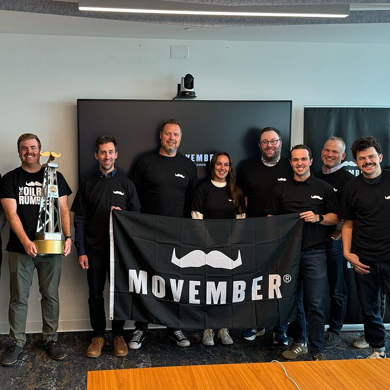 Photo of a group of people, posing for a photo in front of a Movember flag.