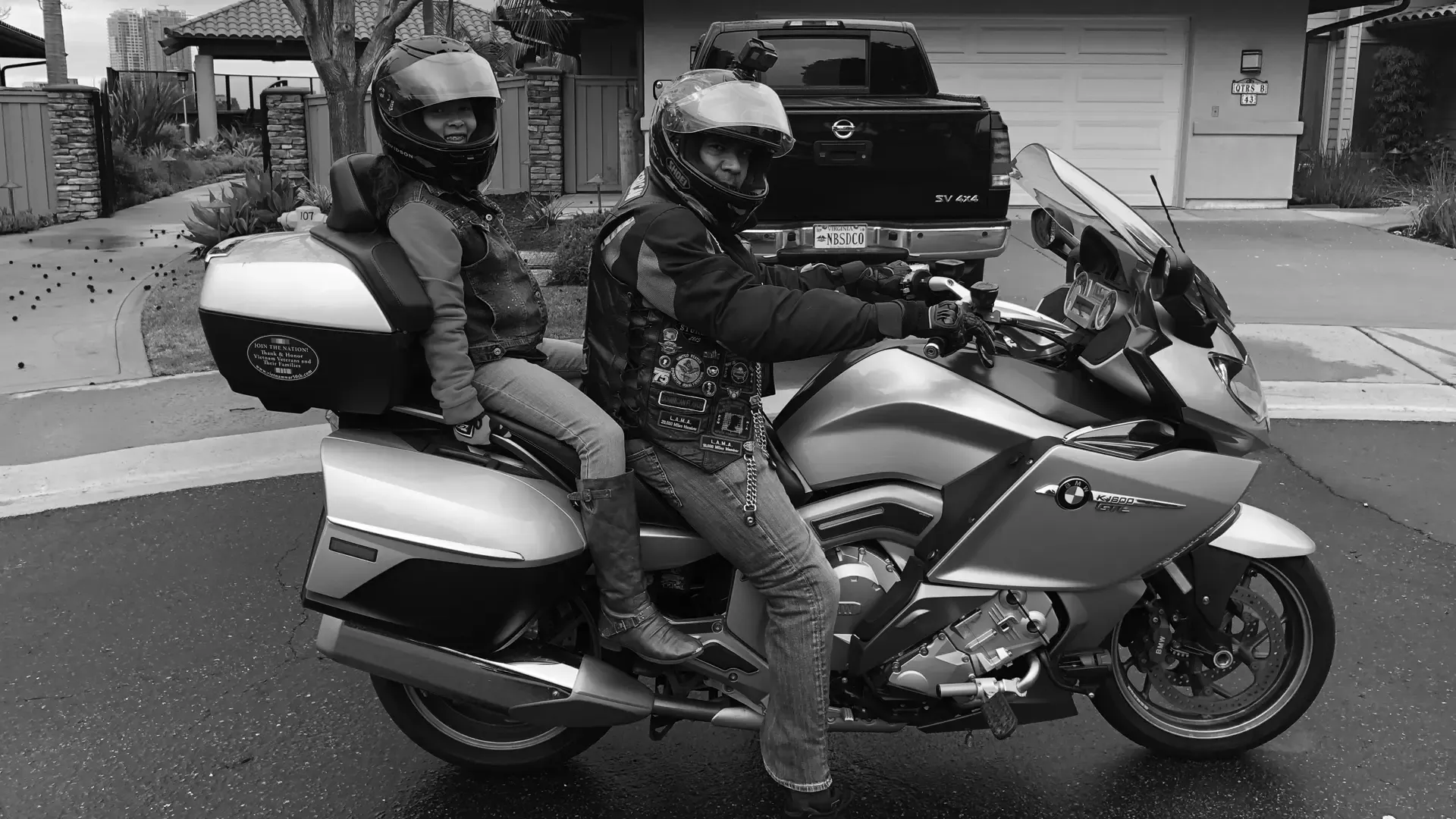 Black and white photo of two people looking sideways to camera, dressed in full gear and sitting on a motorcycle.