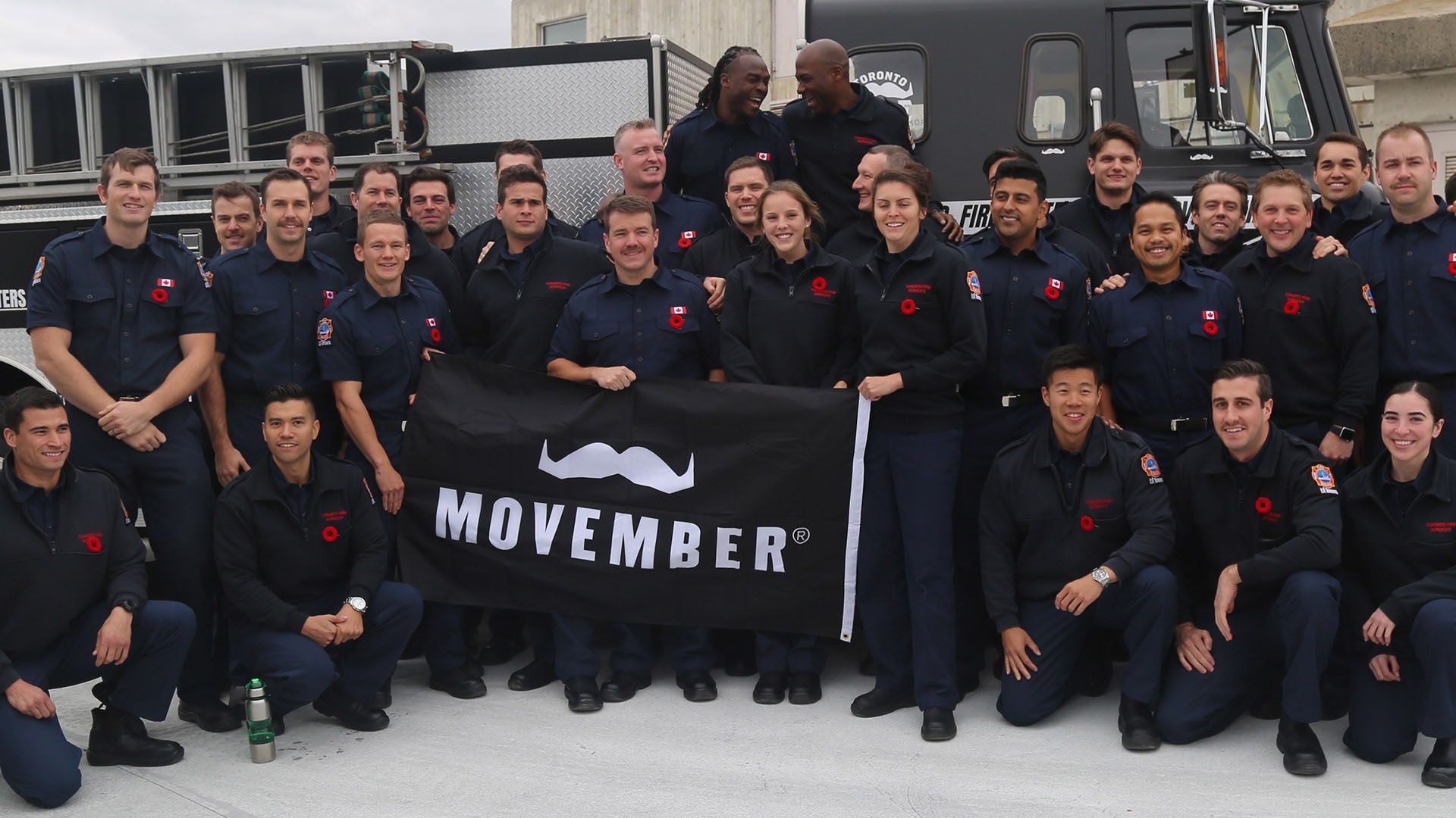 A full fire station crew in uniform, posing to camera while holding a Movember banner.