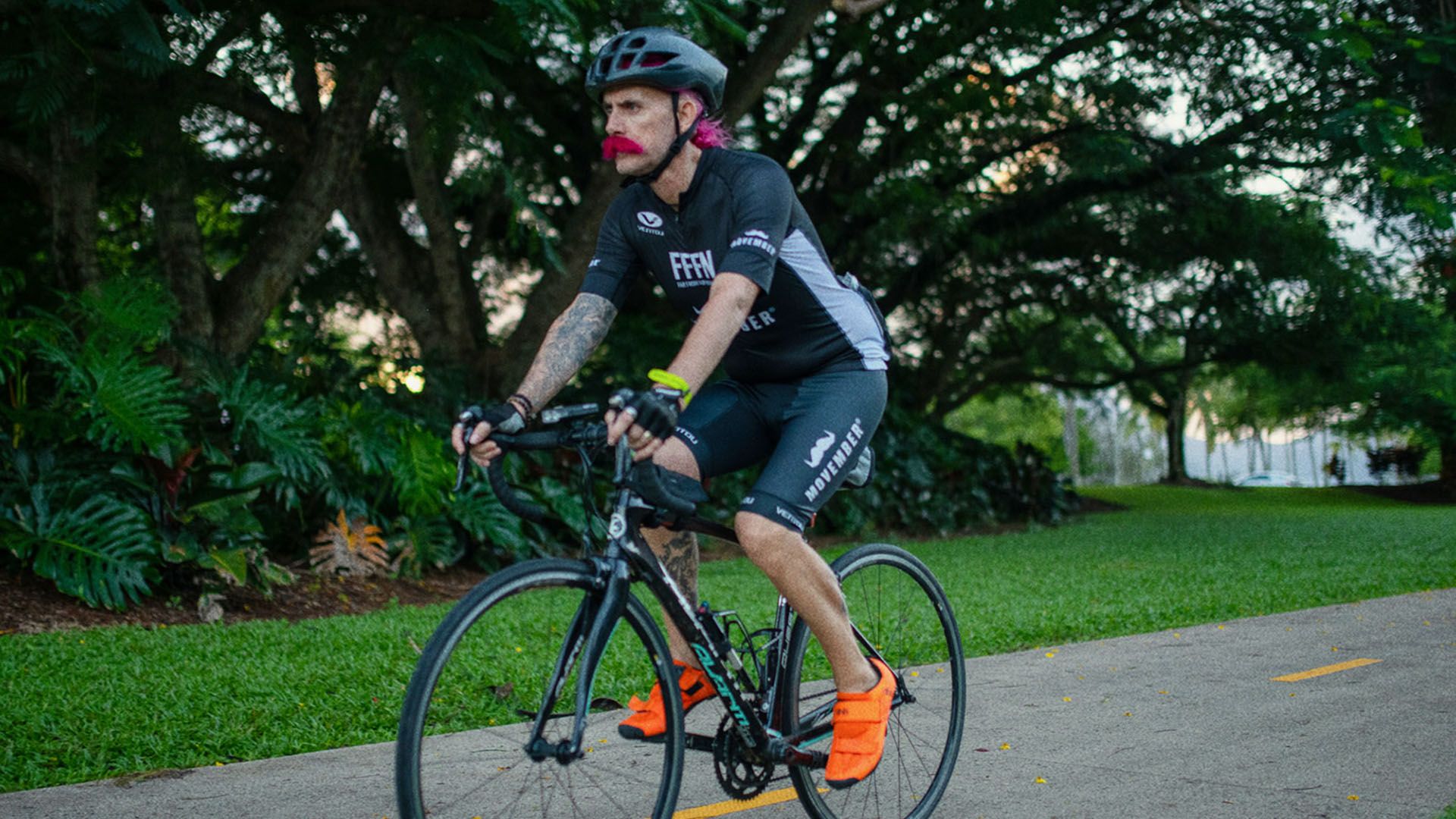 Man with pink moustache rides his bike on a footpath with trees in the background