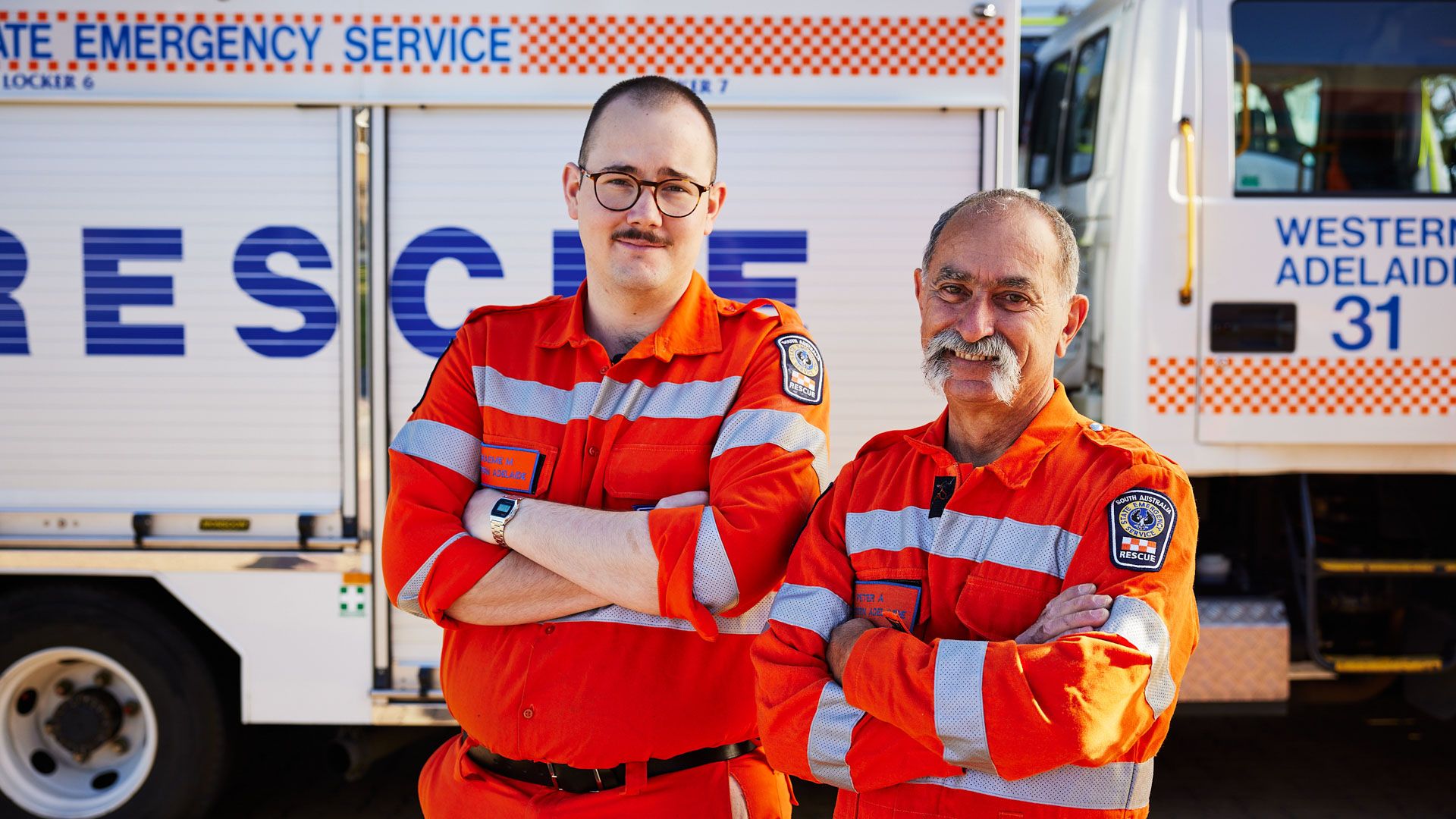 Two emergency service members in orange uniform stand with arms folded and smiling to camera, with their vehicle in the background.