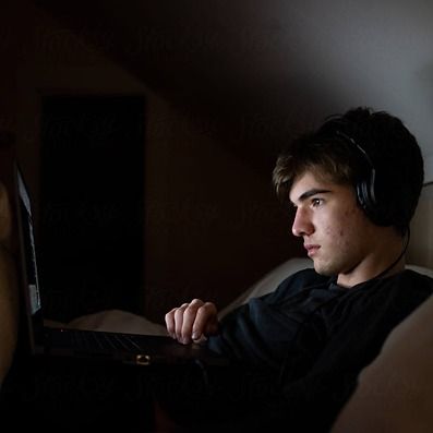 A young man sitting in the dark staring at his laptop screen with headphones on.
