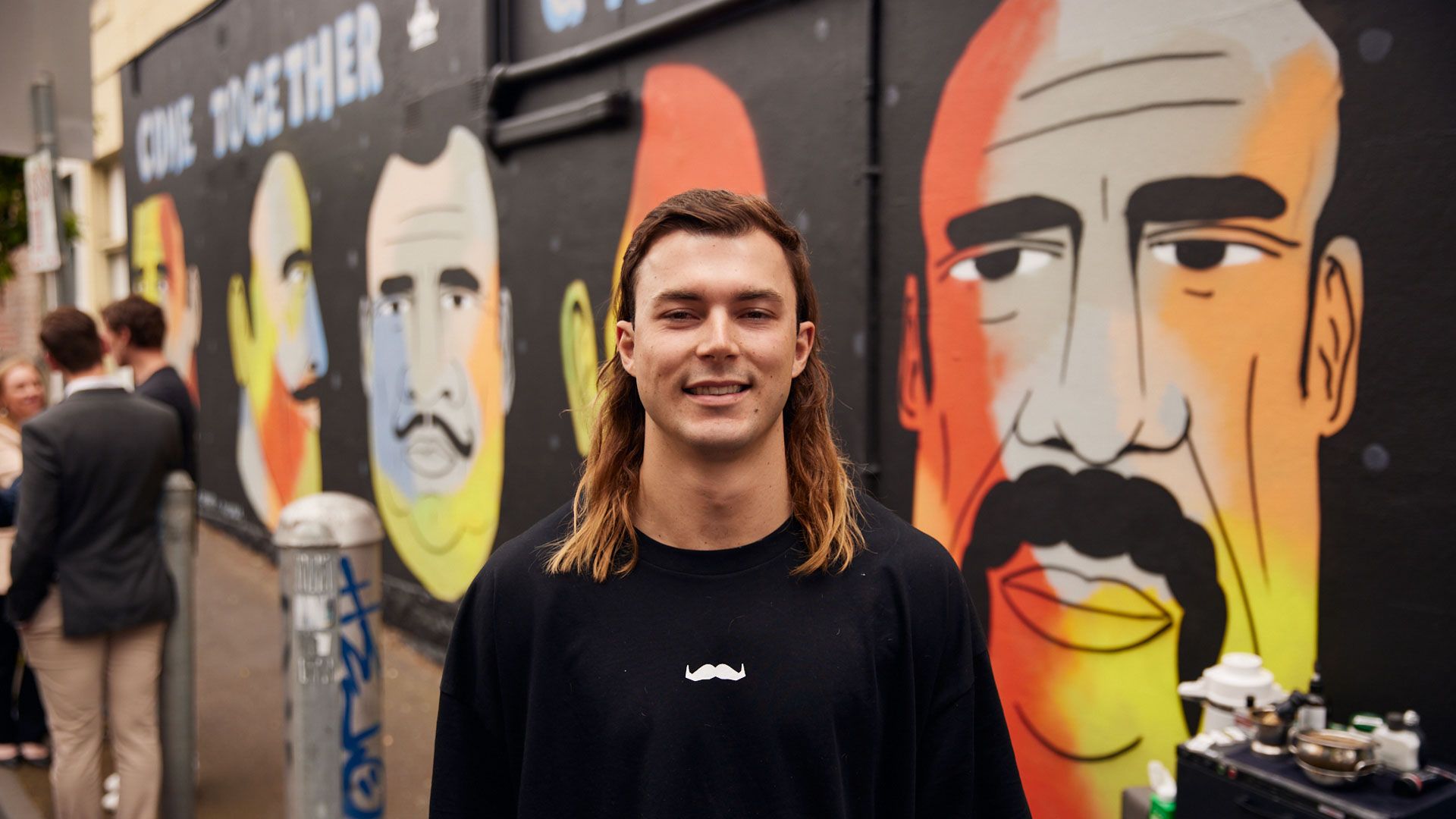 Tall man with long hair stands in front of street art mural and smiles at camera