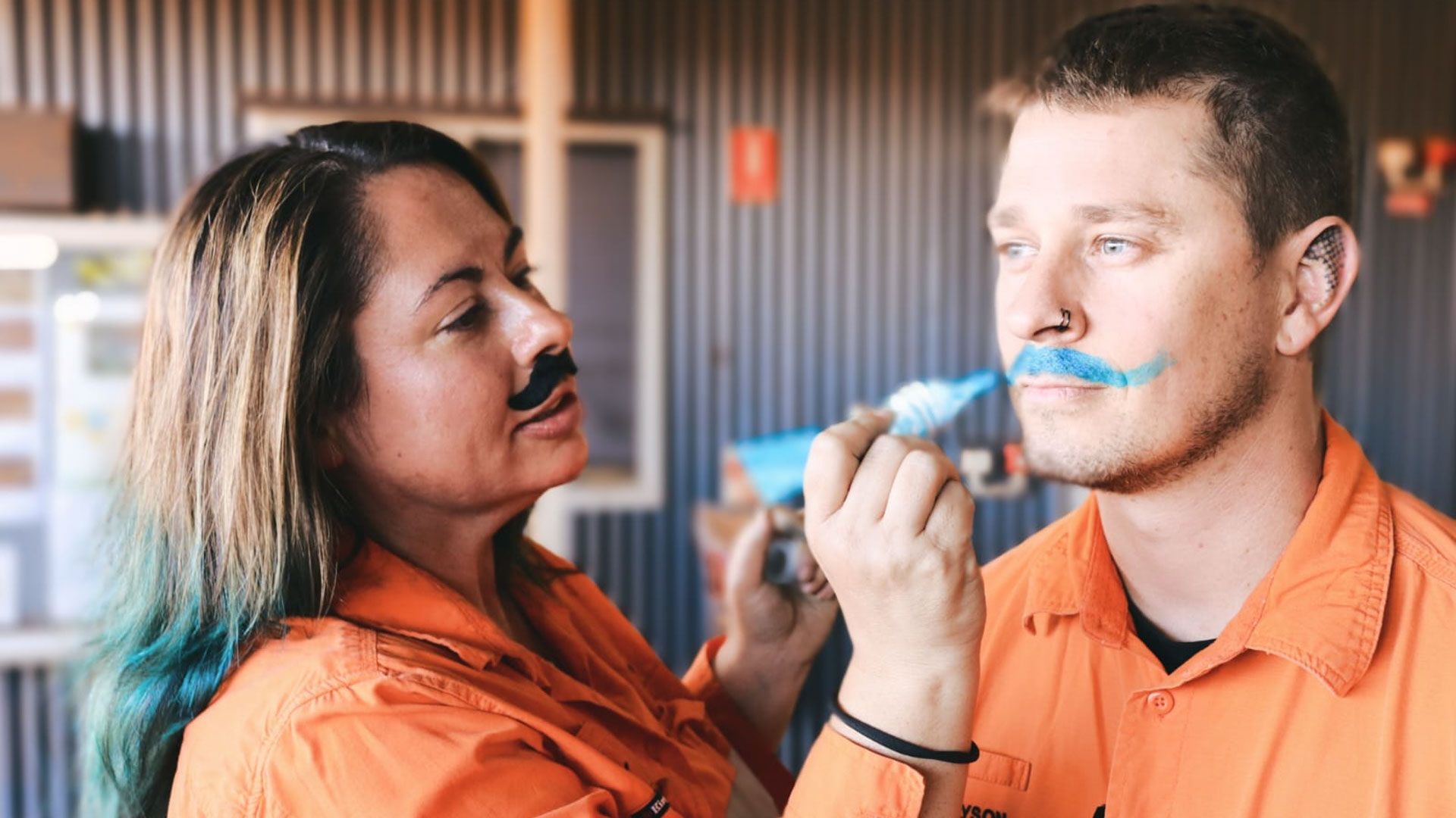 Two people in hi-vis draw moustaches on each others faces