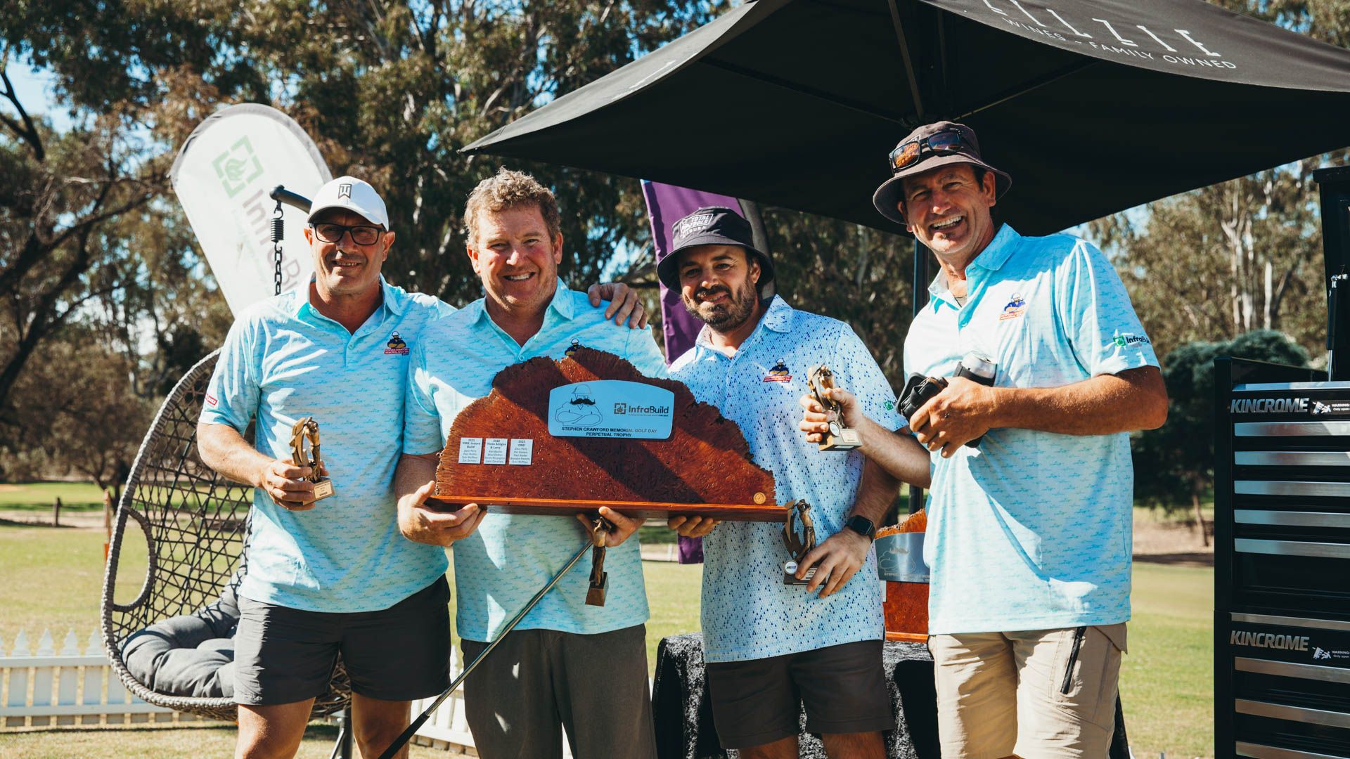 Four men in golf outfits hold trophy in front of tent