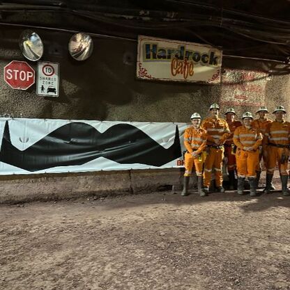 Miners in an underground shaft, posing before a large Movember moustache sign.