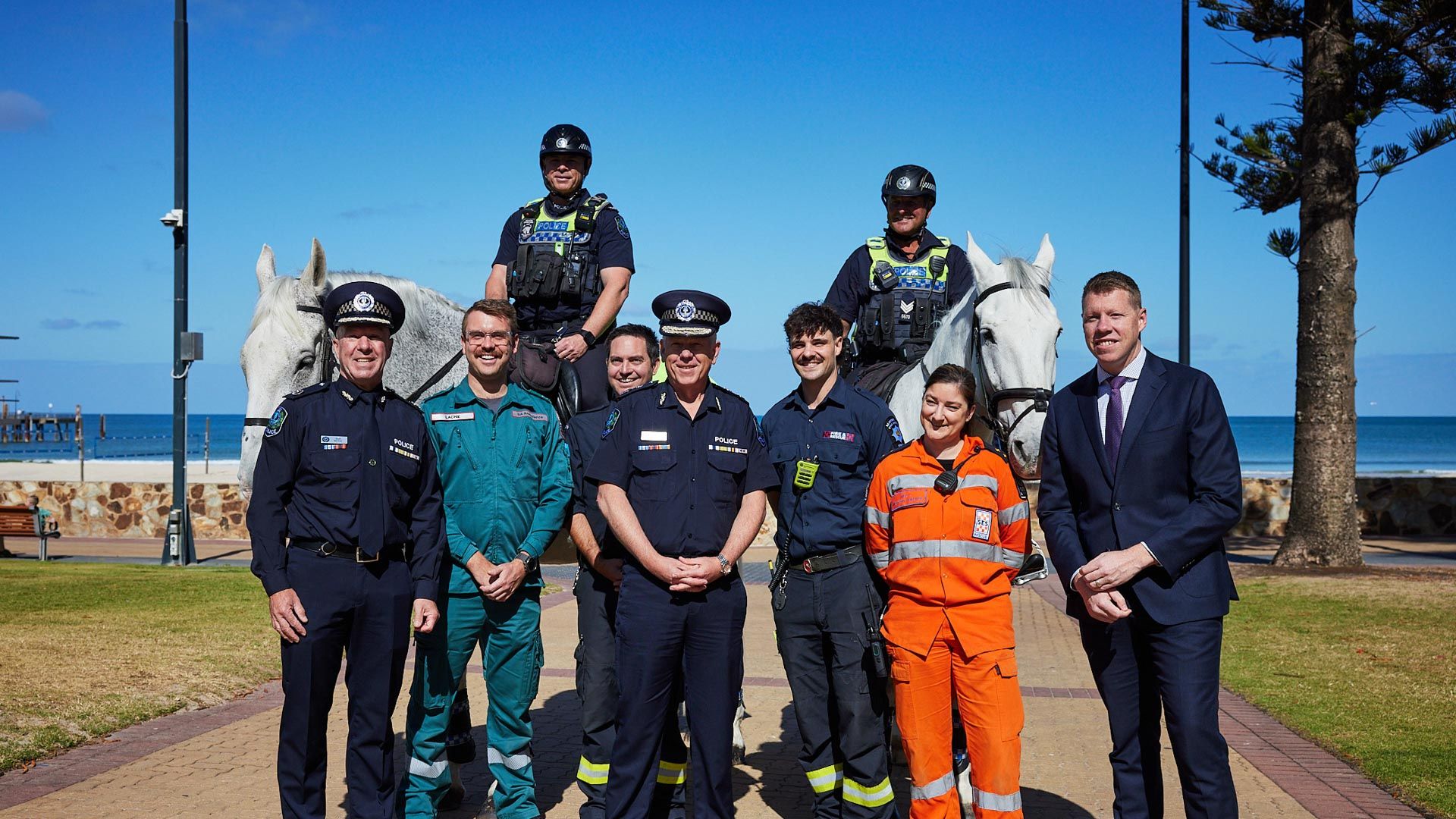 9 people all looking at camera in different emergency service uniforms, with two police men on horseback behind. Blue sky and beach in the background.