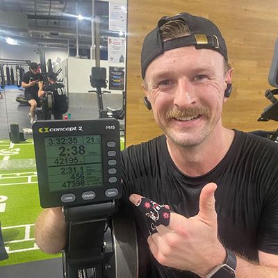 Man sporting a rich moustache pointing to results on a rowing machine's digital display.