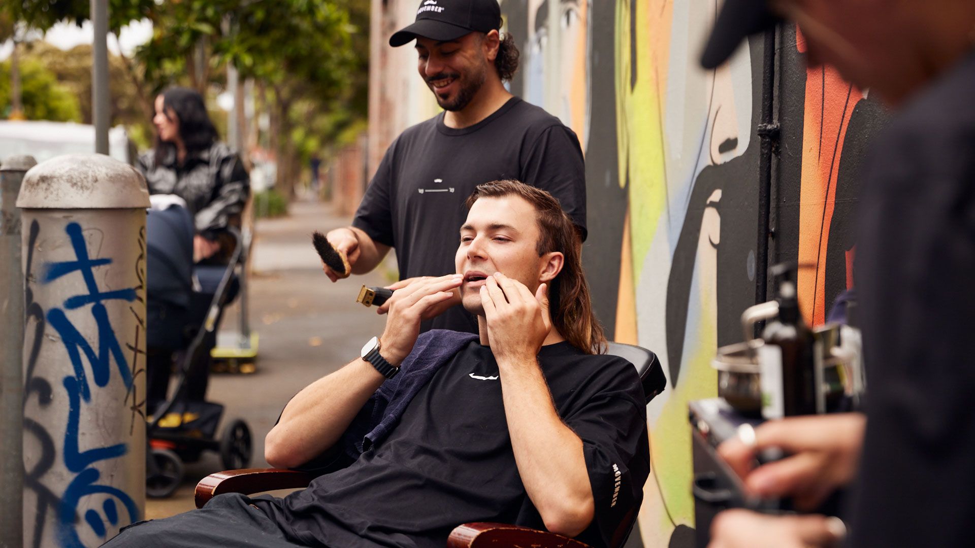 Man sits in barber chair touching his moustache before it is shaved by the barber preparing behind him