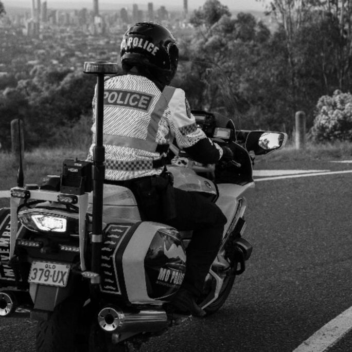 Black and white photo, showing tear, three-quarter shot of a police officer riding a motorcycle. The bike features Movember logos.